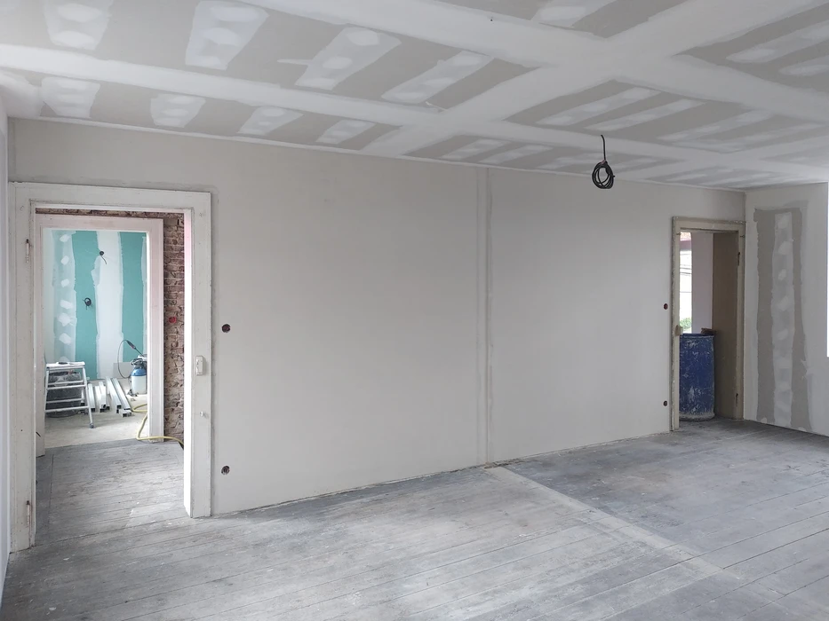 Intérieur en cours de rénovation avec murs en placo brut non peints, plafond avec poutres apparentes blanches