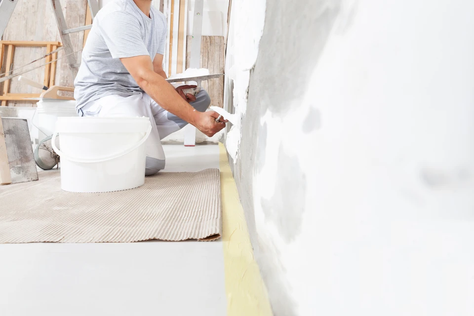 Jeune homme souriant accroupi dans une cuisine moderne blanche, tenant un pinceau plat avec manche en bois et un pot de peinture blanc